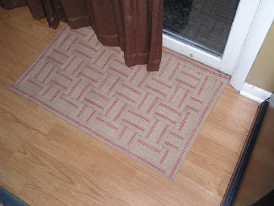 A beige and red patterned upholstery webbing rug doormat is placed on a wooden floor in front of a glass door with brown curtains.