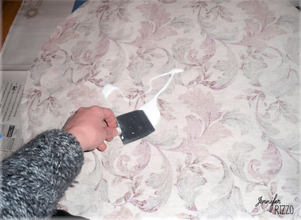 A hand uses a metal scraper to smooth fabric with a pastel floral pattern on a flat surface, preparing it to decoupage a table top.