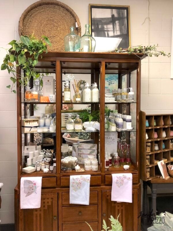 A wooden cabinet displays various bath and body products, decorative jars, towels, and plants, with framed art and baskets on top.