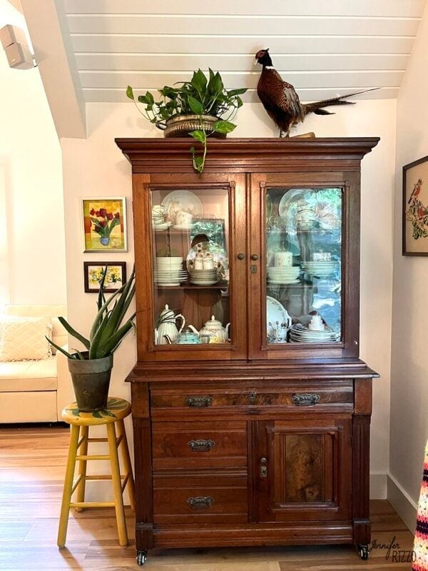 A wooden china cabinet displays dishes behind glass doors, with a potted plant and a pheasant figure on top. A plant in a pot sits on a yellow stool to the left.
