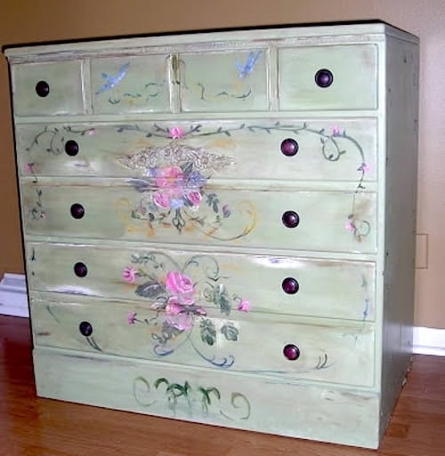 A light green wooden dresser with seven drawers, featuring Ethan Allen Furniture Makeovers touches—hand-painted floral designs and birds—stands on a hardwood floor against a tan wall.