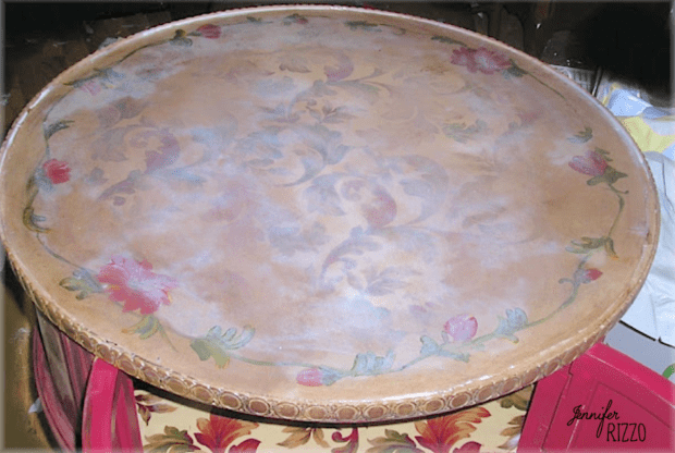 A round wooden tray with a faded floral pattern and decorative edge, reminiscent of the charming style you might use to decoupage a table top, resting on top of a floral-patterned box.