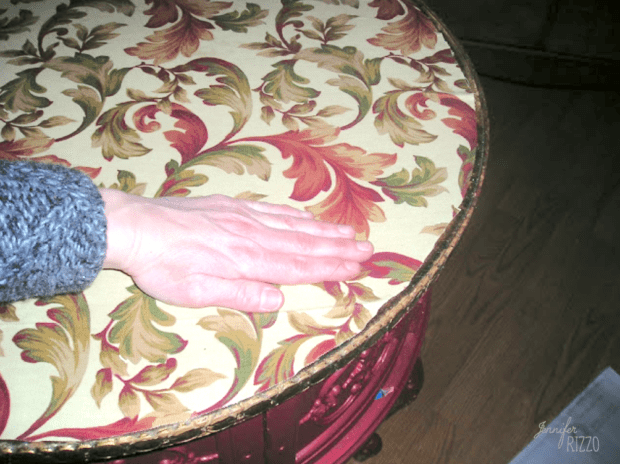 A hand rests on a round table with a floral-patterned top and a decorative red edge, showing how to decoupage a table top for a charming, personalized touch.