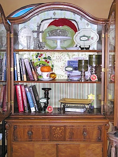 A wooden display cabinet with glass doors contains books, plates, candlesticks, glassware, a pumpkin ornament, and decorative items arranged on three shelves.