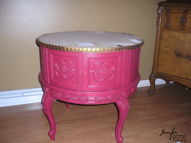 A round, vintage side table painted pink with decorative floral details, missing its tabletop—perfect for those looking to decoupage a table top—stands on wooden flooring near a brown wall and dresser.