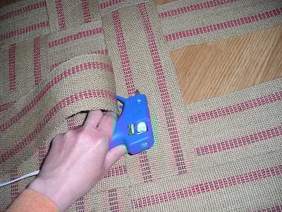 A person uses a blue hot glue gun to attach the edge of an upholstery webbing rug or mat to a wooden surface.