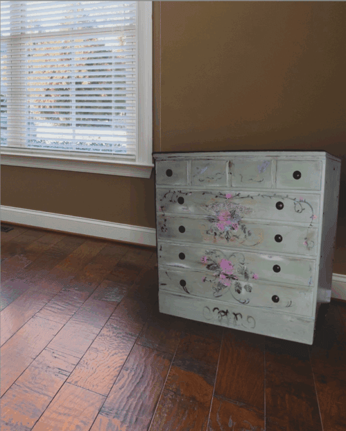 A painted dresser with floral designs, inspired by Ethan Allen Furniture Makeovers, sits on a wooden floor in a room with brown walls and a large window with white blinds.
