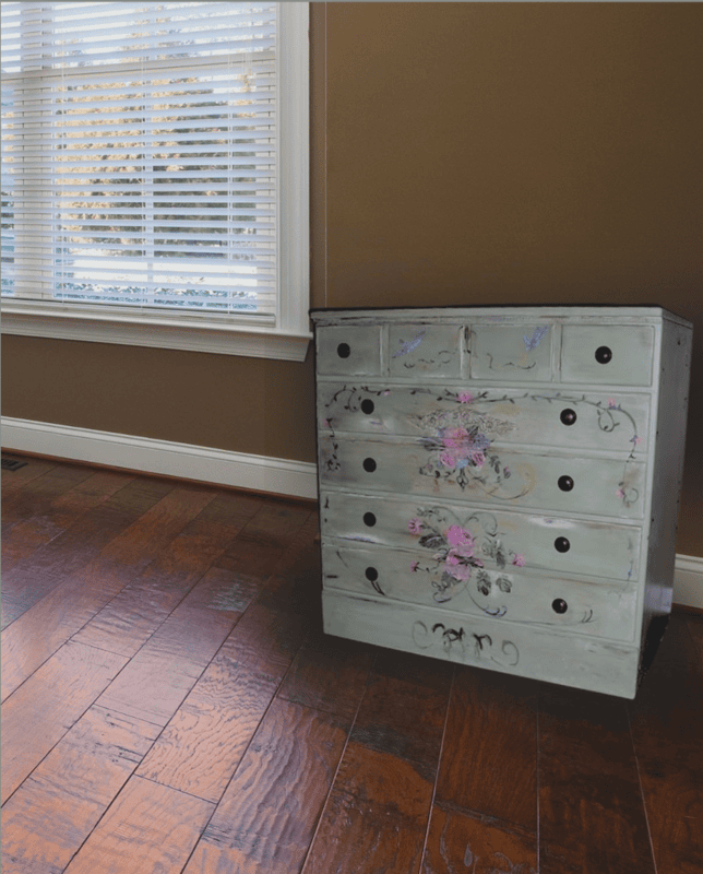 A painted dresser with floral designs, inspired by Ethan Allen Furniture Makeovers, sits on a wooden floor in a room with brown walls and a large window with white blinds.