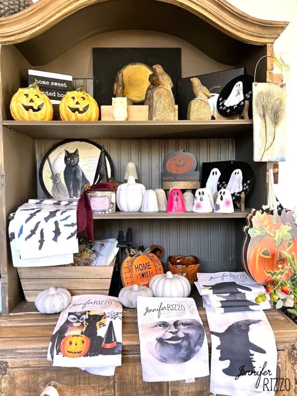A wooden shelf displays Halloween-themed decorations, including pumpkins, ghost figurines, black cat art, and holiday towels, with a “Jennifer Rizzo” label on several items.