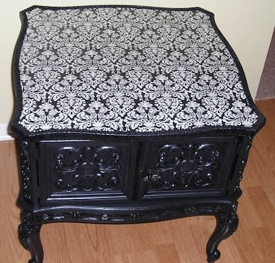 Black vintage side table with ornate carved legs and doors, featuring a stylish side table makeover with a decoupaged table top in black and white damask, placed on a wooden floor against a light-colored wall.