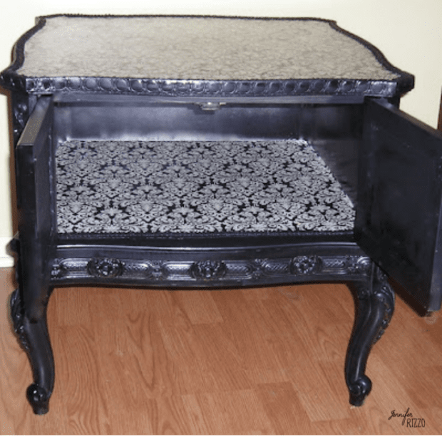Black vintage wooden side table with ornate carvings, open cabinet doors, and a patterned black-and-white interior. This elegant piece showcases a stylish side table makeover with a decoupaged table top, set on a classic wooden floor.