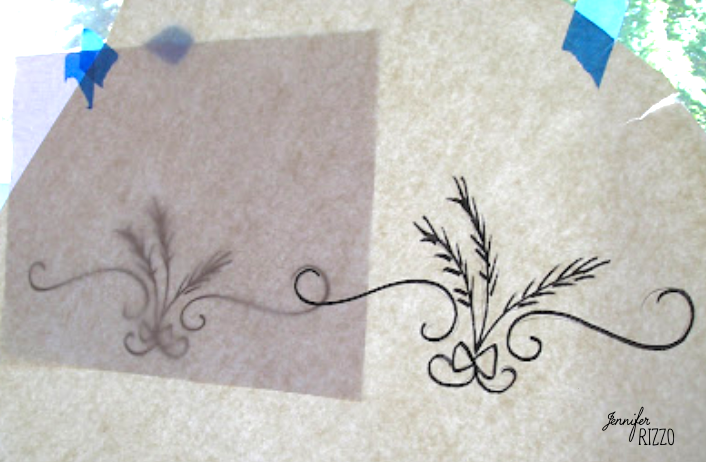 A tracing paper with a floral and ribbon design on the left is placed over a similar completed design on the right, demonstrating how to transfer a repeating image or pattern with a pencil. Blue tape holds the tracing paper at the top.