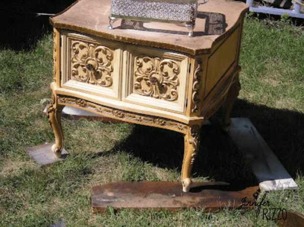 Ornate, vintage wooden side table with carved floral designs on the doors, featuring a charming side table makeover with a decoupaged table top, standing on grass with some boards underneath its legs.