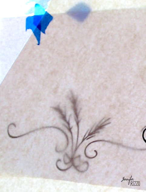 Image of a delicate, dark stencil design with swirls and branches on a light background. Blue tape is visible at the top. The corner of a white paper is slightly covering the stencil, showing how to transfer a repeating image or pattern with a pencil.