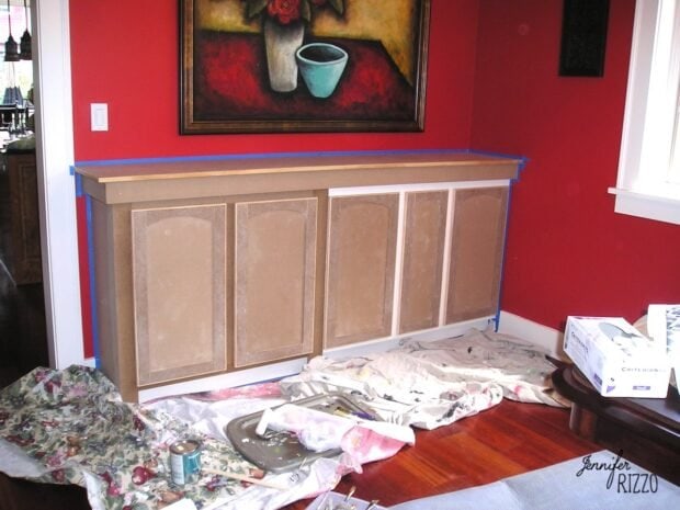 A room with red walls features an arts and crafts inspired painted built in cabinet, with drop cloths, paint cans, and supplies on the floor, and blue painter’s tape outlining the cabinet edges.