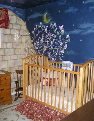 A wooden crib sits in a nursery with a baby nursery wall mural of a night sky, crescent moon, and flowering tree painted on the wall. A dresser and rocking chair are nearby.