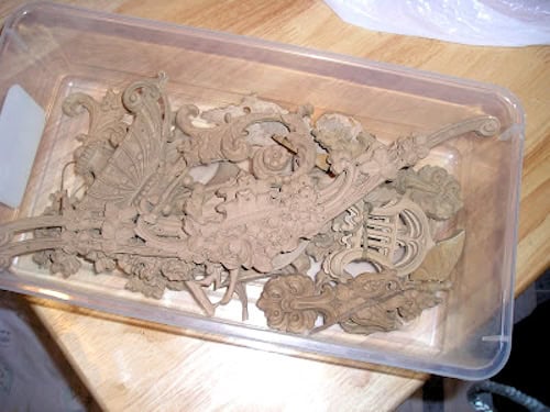 A clear plastic container filled with intricately carved, unfinished wood embellishments sits on a wooden table.