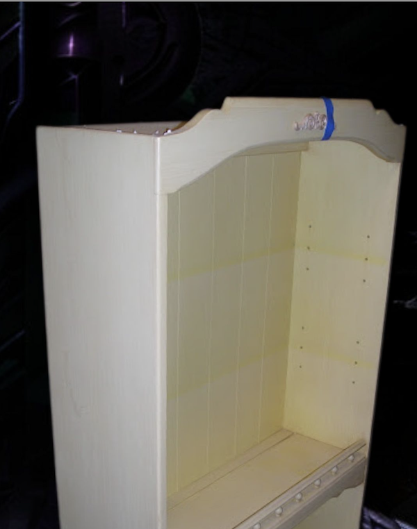 A light-colored, unfinished wooden shelf or cabinet with a curved top and tasteful wood embellishments, set against a dark background.