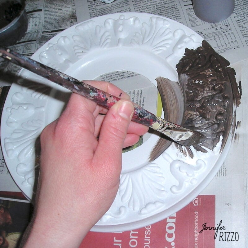 A hand holds a paintbrush, painting brown paint onto the edge of a white decorative ceiling medallion to look old resting on newspaper.