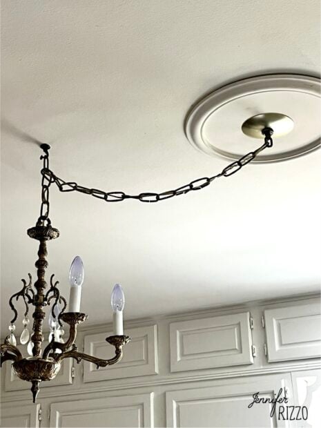 A brass chandelier hangs from a ceiling medallion, suspended by a long chain that is attached off-center. White cabinets are visible below.