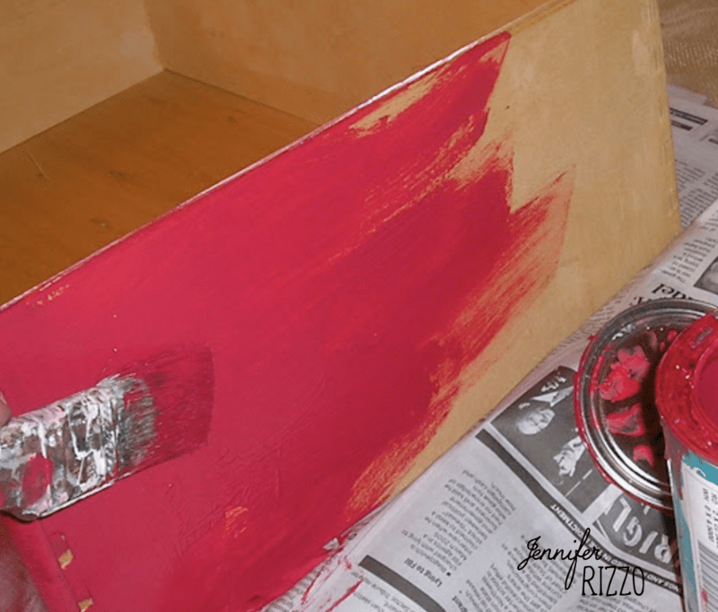 A wooden drawer, repurposed from a dresser into a planter, is being painted red with a paintbrush; a can of red paint and newspapers are nearby.