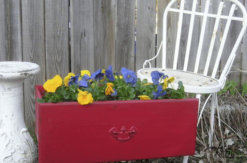 A repurposed dresser drawer into a planter, painted red and filled with yellow and purple flowers, sits next to a white metal chair and a white pedestal by a wooden fence.