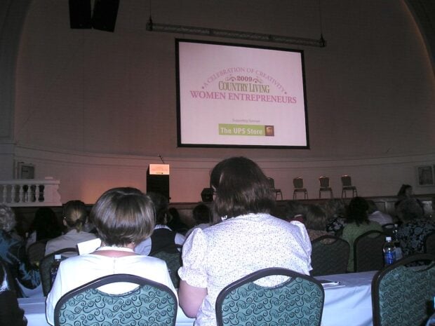 Audience seated in a conference room faces a stage with a projected sign reading "Country Living Women Entrepreneurs 2008," at a creative business conference with Country Living Magazine, sponsored by The UPS Store.