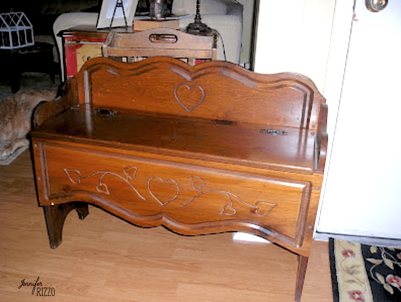 A charming piece of carved wood furniture, this wooden storage bench features a hinged lid and decorative carved hearts on the backrest and front panel, placed on a wooden floor near a white door.
