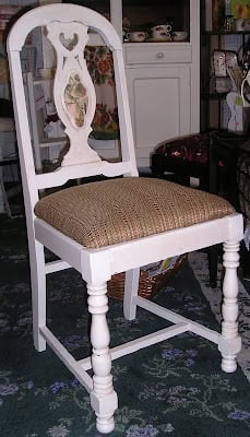 White wooden chair with a carved backrest and a padded, patterned seat cushion, featuring a creative chair makeover with paint and scrapbook paper, placed on a green patterned rug in a room with various decor items.