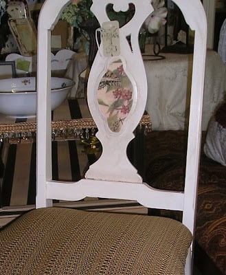 A close-up of a white wooden chair, transformed with a chair makeover using paint and scrapbook paper. It features a decorative backrest and a woven light brown seat cushion, with various decorative items and a bowl in the background.