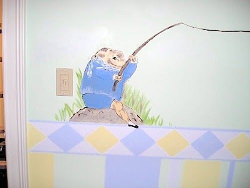 A mural of a small animal in a blue coat fishing, painted on a wall above a row of pastel yellow and blue diamonds. An electrical outlet is nearby.