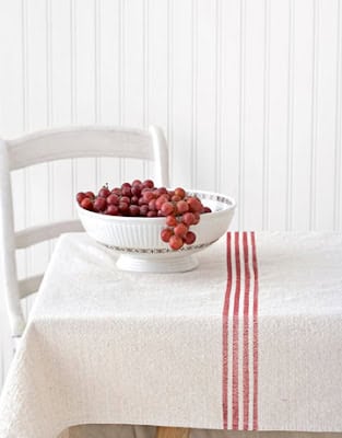 A white bowl filled with red grapes sits on a table covered with a white cloth featuring three red stripes. A white chair is positioned beside the table.