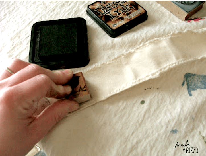 A hand holds a rubber stamp and presses it onto a piece of off-white fabric, creating a hand stamped muslin ribbon, with ink pads visible nearby on the surface.