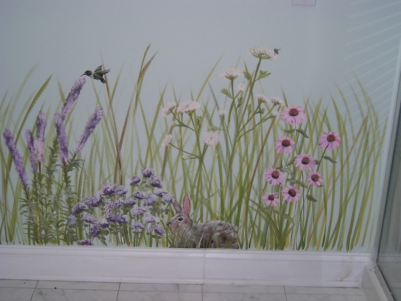 A wall mural inspired by hand painted and stenciled furniture ideas depicts wildflowers, grasses, a rabbit, a hummingbird, and a bee against a pale background above a white tile floor and baseboard.