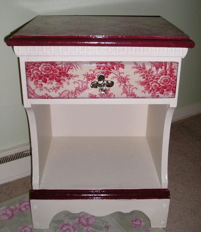 A white wooden nightstand with a single drawer featuring a red floral pattern and a metal handle, a red top and bottom trim, and an open lower shelf.