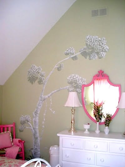 A bedroom corner with a pale green wall featuring a painted tree mural, a white dresser with hand painted and stenciled furniture ideas, a pink-framed mirror, lamps, and floral decorations.