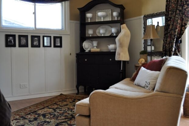 A living room with a beige sofa, a black cabinet with white ceramics, a dress form, framed photos on a Board and Batten wall treatment, and a patterned rug.