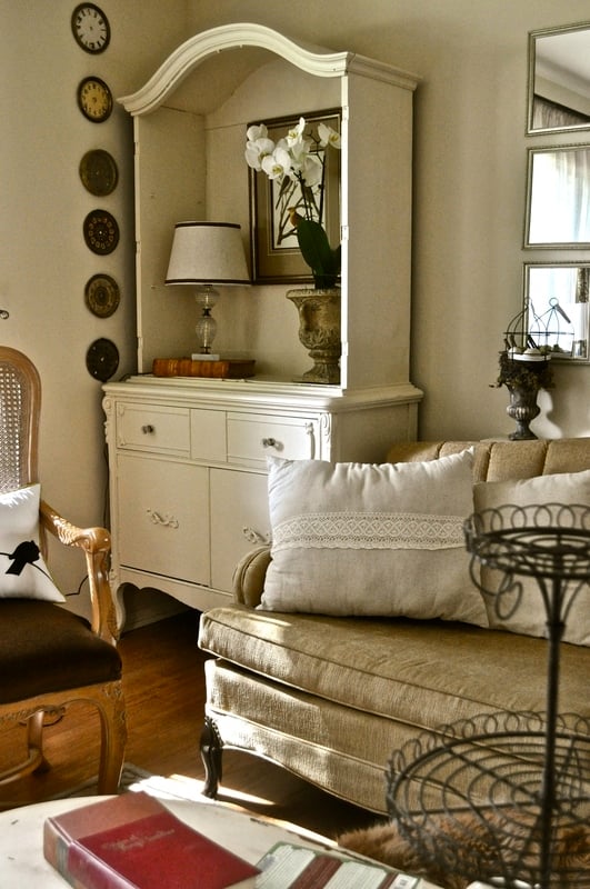 A living room with a beige sofa, white china cabinet turned into a display case, table lamp, potted orchid, wall clocks, and framed artwork. Warm lighting enhances the cozy, elegant decor.