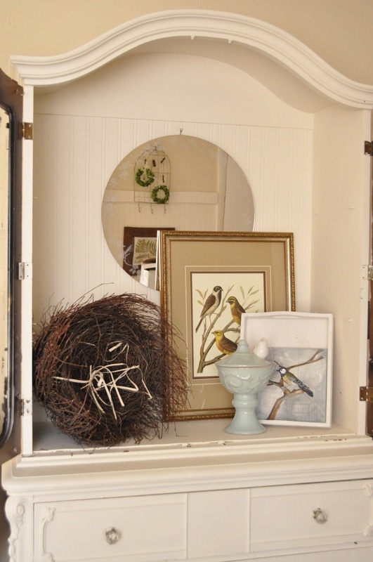White china cabinet turned into a display case with a round mirror, bird illustrations in frames, a large twig nest, and a blue vase holding a ceramic bird and egg on top of the white dresser.