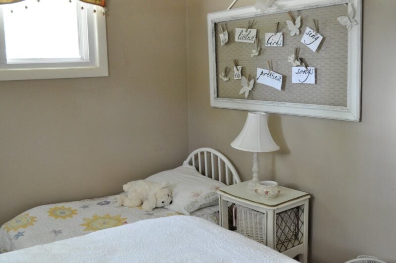 A cozy butterfly-inspired girl's bedroom corner with a single bed, white bedding, a stuffed polar bear, wicker nightstand, lamp, and a decorative frame featuring butterflies and handwritten notes on the wall.