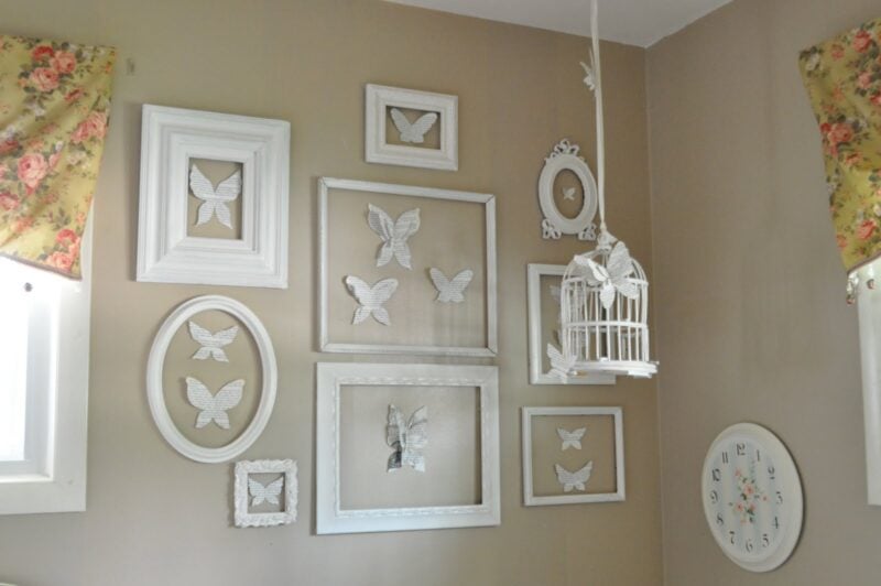A beige wall in a butterfly-inspired girl's bedroom features white-framed butterfly art, a white birdcage, floral curtains, and a round clock with black numbers.