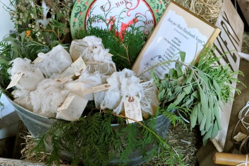A metal tub filled with wrapped soaps, herbs, and foliage, accompanied by handwritten tags and a printed recipe or instructions on paper in the background.