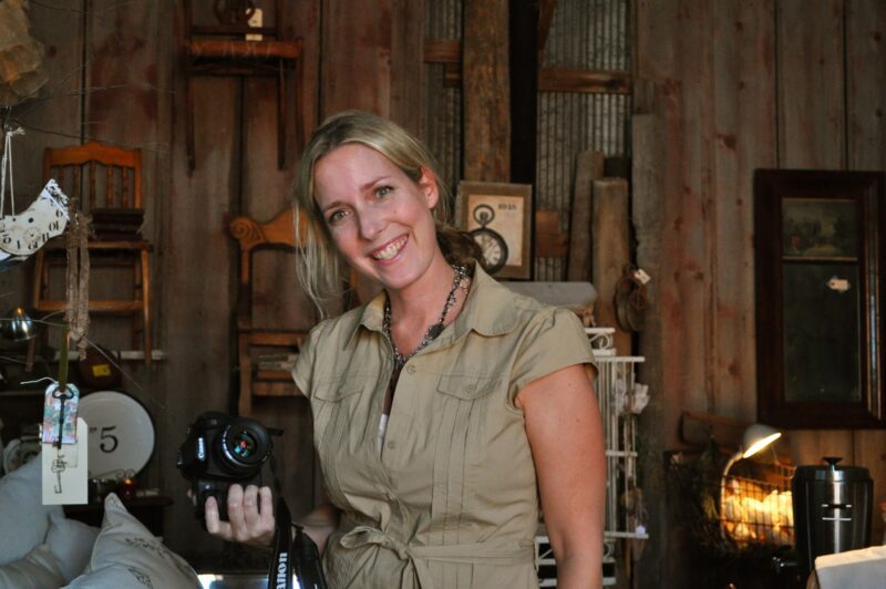 A woman in a beige shirt stands indoors, smiling at the camera and holding a DSLR camera in her right hand. The background features wooden walls and various vintage objects.