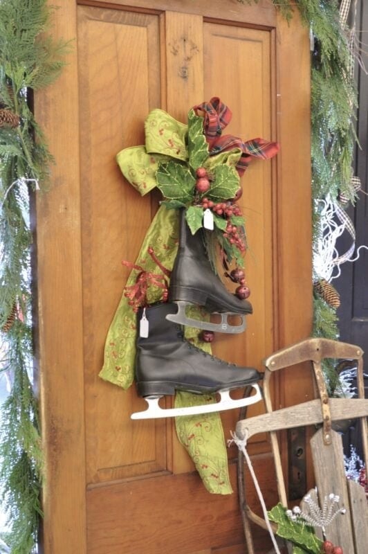 A pair of black ice skates hangs on a wooden door, decorated with a green ribbon and red berries, with greenery and a wooden sled nearby.