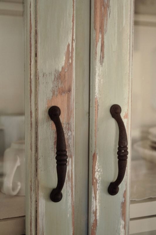 Close-up of two distressed wooden cabinet doors with dark metal handles; chipped paint reveals wood and an aged appearance—perfect kitchen decorating details. White dishes are visible inside the cabinet.