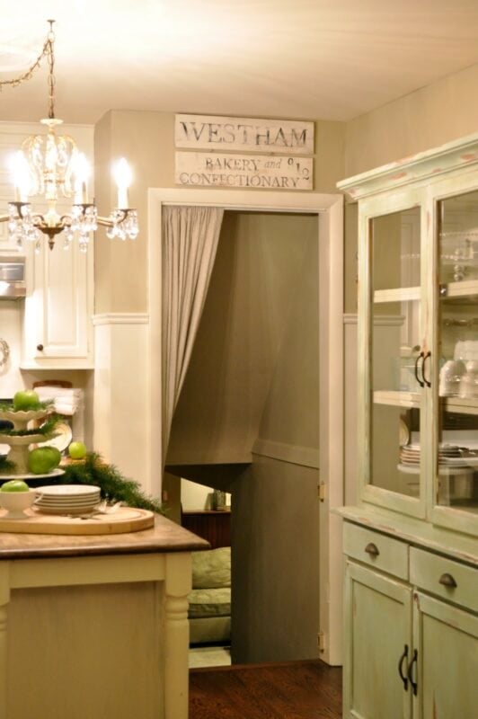A cozy kitchen featuring charming kitchen decorating details—a chandelier, green cabinet showcasing dishware, and a doorway with a curtain and vintage "Westham Bakery and Confectionary" sign above.