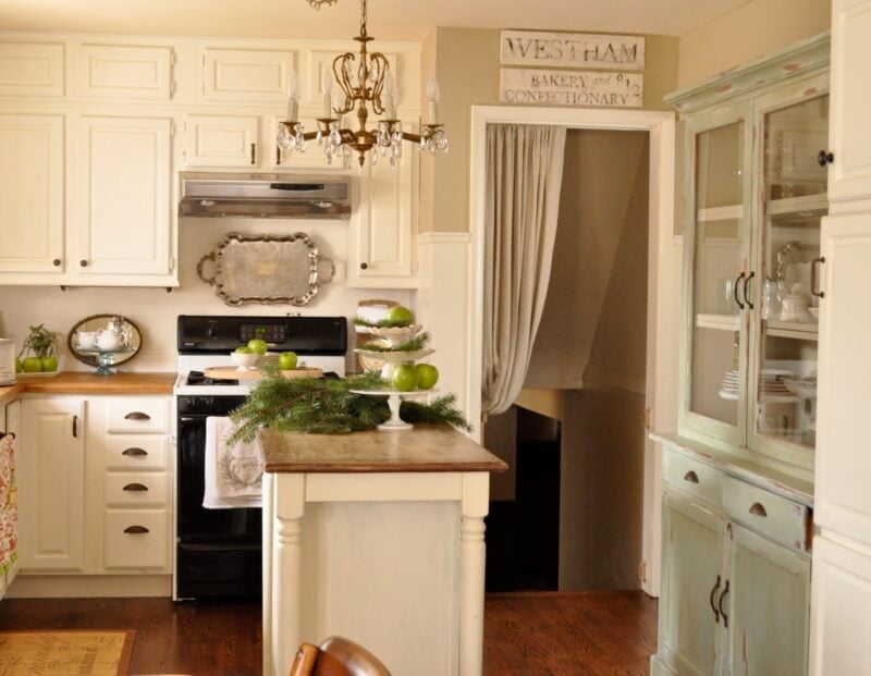 A cozy kitchen with cream cabinets and kitchen decorating details like a wood-topped island adorned with green apples and pine branches, a stove, a mint green hutch, and a chandelier.