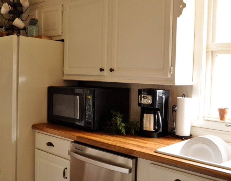 A kitchen countertop with a microwave, coffee maker, paper towel roll, and a small plant showcases charming kitchen decorating details beneath white cabinets near a refrigerator and a window.