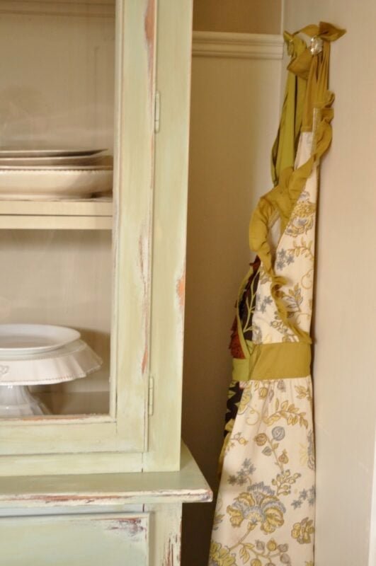 A floral apron with green straps hangs on a hook beside a pale green vintage cabinet displaying white dishes, adding charming kitchen decorating details.