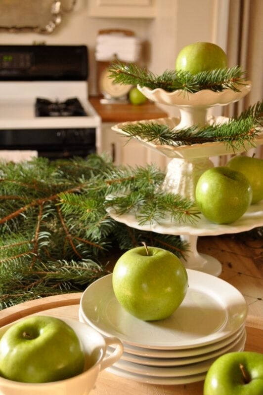 Green apples and pine branches arranged on a tiered stand and white plates add charming kitchen decorating details, with a stove in the background.